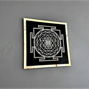Puede incluir: Un cuadrado negro con marco dorado, que presenta un dise&ntilde;o blanco de Sri Yantra. El Sri Yantra es un patr&oacute;n geom&eacute;trico complejo utilizado en el hinduismo y otras tradiciones.
