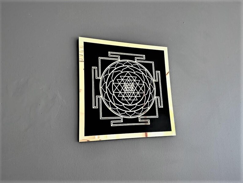Puede incluir: Un cuadrado negro con marco dorado, que presenta un dise&ntilde;o blanco de Sri Yantra. El Sri Yantra es un patr&oacute;n geom&eacute;trico complejo utilizado en el hinduismo y otras tradiciones.