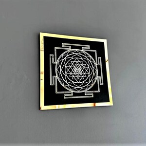 Puede incluir: Un cuadrado negro con marco dorado, que presenta un s&iacute;mbolo Sri Yantra blanco en el centro. El s&iacute;mbolo es un dise&ntilde;o geom&eacute;trico con tri&aacute;ngulos y c&iacute;rculos.