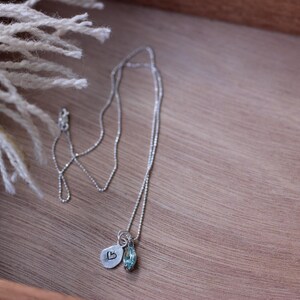 May include: A silver necklace with two pendants rests on a wooden surface. One pendant is a small, stamped metal charm with a heart, and the other is a blue gemstone. A white fringed textile is in the upper left corner.