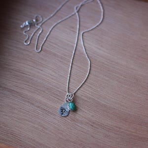 May include: A silver necklace with a small pendant and a turquoise stone. The pendant has a stamped design. The necklace is displayed on a wooden surface.