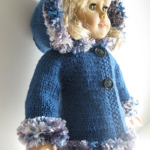 Puede incluir: Un abrigo de punto azul para muñeca con un ribete de pelo blanco y gris alrededor de la capucha y los puños. El abrigo tiene dos botones negros.