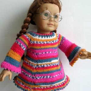 May include: A doll wearing a colorful striped knitted sweater dress with bell sleeves. The doll has brown hair and is wearing round glasses.