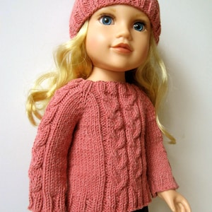 May include: A doll wearing a pink knitted sweater with a cable knit pattern and a pink knitted hat. The doll has blonde hair and blue eyes.