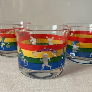Könnte beinhalten: Drei transparente Trinkgläser mit einem Regenbogen-Design. Jedes Glas hat ein Band aus Rot, Orange, Gelb, Grün und Blau mit Silhouetten von Kindern, die Sport treiben. Der Text "Chicago Park Kids" ist ebenfalls sichtbar.