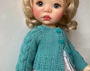 May include: A teal knitted cardigan with a cable knit pattern and button closure. The cardigan is worn by a doll with blonde hair and green eyes.