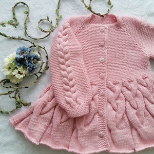Peut inclure: Un cardigan rose en tricot avec un motif en câble et un ourlet à volants. Le cardigan a une fermeture à boutons et est fait avec un fil doux.