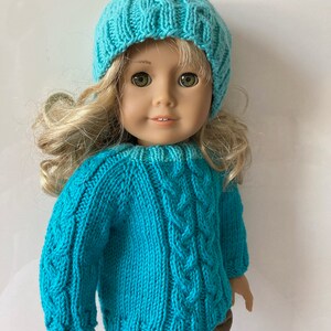 May include: A turquoise knitted sweater with a cable knit pattern and matching turquoise knit hat. The sweater and hat are worn by a doll with blonde hair.