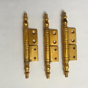 May include: Three ornate brass hinges with decorative finials. Each hinge has a rectangular base with two screw holes and a long, vertical section with a detailed design. The hinges are a warm, golden color.