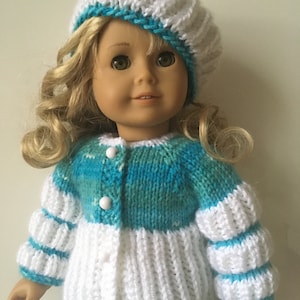 Könnte beinhalten: Ein gestrickter Puppencardigan mit weißen und blauen Streifen. Der Cardigan hat einen Knopfverschluss und Puffärmel. Die Puppe trägt eine weiße und blaue Strickmütze.