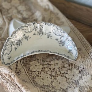 May include: A crescent-shaped ceramic bone dish featuring a white base with a detailed grey floral pattern and decorative scrollwork along the scalloped edges. The dish has a curved design and a smooth, glossy finish with visible intricate botanical motifs.
