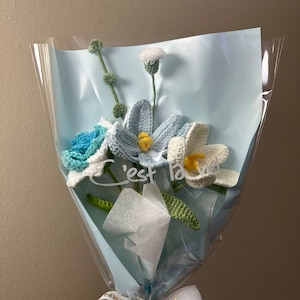 May include: A bouquet of crocheted flowers in shades of blue, white, and green, wrapped in a light blue cellophane with a white tissue paper liner. The bouquet is tied with a white ribbon with gold lettering that reads "Happiness is Eternal". The flowers are arranged in a vase and the text "C'est la vie" is written on the cellophane.