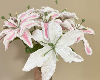 May include: A bouquet of crocheted white lilies with pink accents in a clear glass vase. The lilies have green stems and leaves.