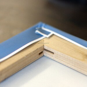 May include: Close-up of a wooden canvas frame with blue fabric stretched across it. The frame is made of two pieces of wood joined together at a corner. The fabric is stapled to the frame.