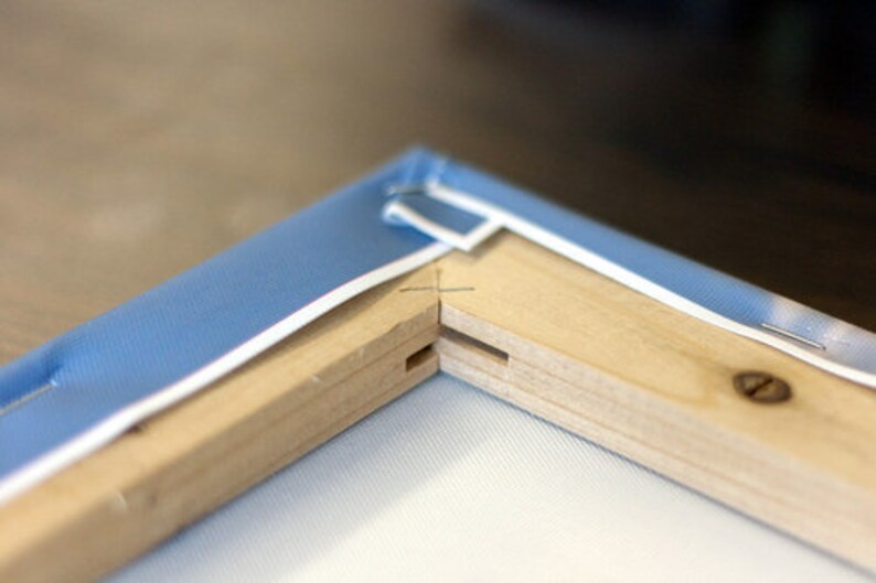 May include: Close-up of a wooden canvas frame with blue fabric stretched across it. The frame is made of two pieces of wood joined together at a corner. The fabric is stapled to the frame.