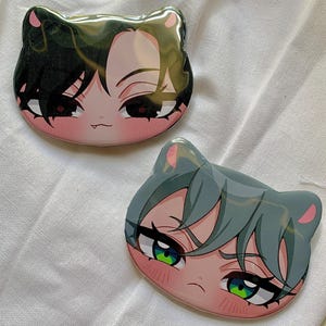 May include: Two cat-shaped pins featuring anime-style character faces. One pin has black hair and red eyes, the other has gray hair and green eyes. Both pins have pink accents and a glossy finish.
