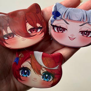 May include: Three cat-shaped anime-style character pins. The pins feature detailed facial expressions with large eyes and colorful hair. The top left pin has red hair and eyes, the top right has silver hair and pink eyes, and the bottom pin has red and white hair with blue eyes.