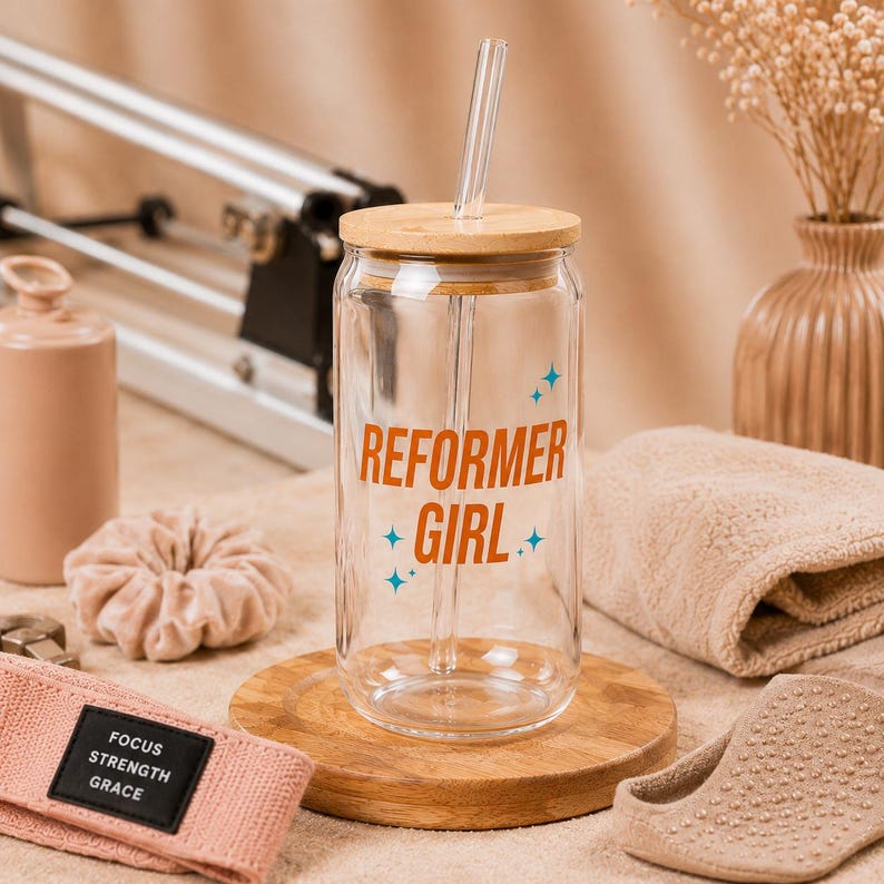 以下が含まれることがあります： 竹製の蓋とガラスストローが付いた透明なガラスタンブラー。「REFORMER GIRL」の文字がオレンジ色で、青い星のアクセントが付いています。タンブラーは木製のコースターの上に置かれ、ピンクのヘッドバンドやその他のアクセサリーが添えられています。