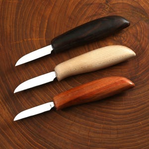 May include: Three carving knives with wooden handles in varying colours: dark brown, light tan, and reddish-brown. Each knife has a silver blade and a curved handle, displayed on a wooden surface.