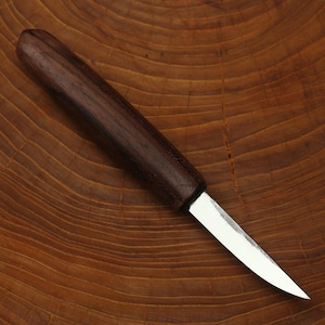 May include: A handcrafted knife with a dark brown wooden handle and a silver blade. The knife is set against a wooden background, showcasing the natural grain of the wood. The blade is sharp and pointed, suggesting a tool for precision cutting.