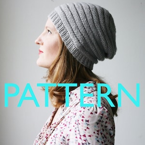 May include: A gray knitted beanie with a ribbed texture. The beanie is worn by a person with long, wavy, light brown hair. The word "PATTERN" is written in large, turquoise letters across the bottom of the image.