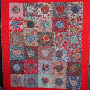 May include: A red quilt with a patchwork design featuring various floral patterns and hearts. The quilt is made up of 25 squares, each with a different fabric. The quilt is edged with a red border.