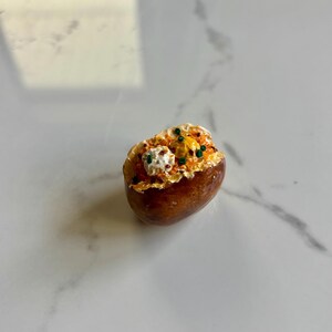 May include: A small, detailed model of a baked potato. The potato has a dark brown skin and is topped with a mixture of white, yellow, and green ingredients, possibly cheese, butter, and herbs. This miniature food item could be a decorative piece.
