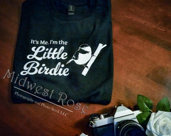 May include: Black t-shirt featuring the text "It's Me. I'm the Little Birdie" and a white bird illustration. The shirt is folded on a wooden surface. A camera and white roses are in the background.