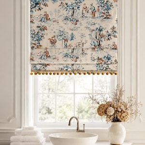 May include: A Roman shade featuring a cream-colored fabric with a blue and rust-colored toile print depicting pastoral scenes of people and trees. The bottom edge of the shade is finished with a decorative gold trim featuring small pom-poms.