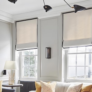 May include: Three white roman blinds with black trim hang in a living room with a white sofa and yellow throw cushions. The blinds are raised to reveal windows with white trim.