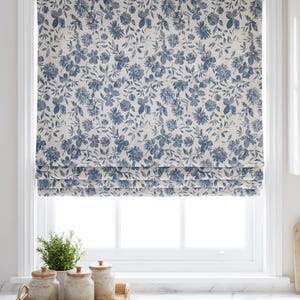 May include: A roman shade featuring a blue floral pattern on a cream background, installed within a white window frame. The fabric displays a dense arrangement of various flowers and leaves, with horizontal folds visible at the bottom of the shade.