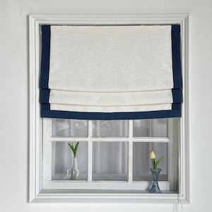 Puede incluir: Estor romano blanco con ribete azul marino. El estor está subido y la ventana que hay detrás tiene un marco blanco y seis cristales. Hay dos pequeñas plantas en maceta en el alféizar de la ventana.