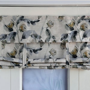 May include: A Roman blind with a floral pattern in beige, grey, and olive green. The fabric displays a botanical design of leaves and flowers. The blind is mounted on a white door frame, providing a decorative window treatment.