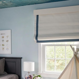 Könnte beinhalten: Ein weißer Rollladen mit blauem Besatz hängt an einem Fenster. Der Rollladen ist hochgezogen und gibt den Blick auf Bäume im Freien frei. Ein weißes Tipi-Zelt steht im Raum.