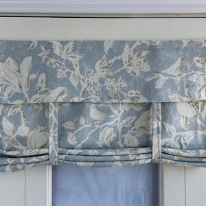 Puede incluir: Una cortina romana de patrón floral azul y blanco con un ribete blanco. La cortina está subida hasta la parte superior de la ventana, dejando ver un marco de ventana blanco.