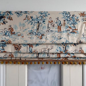 以下が含まれることがあります： 森の風景の中に人々や動物が描かれた、青と茶色の模様のクリーム色のローマシェード。シェードには金色のタッセルが付いている。