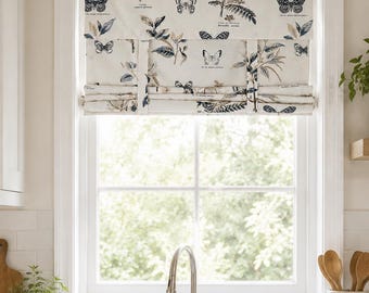 May include: A cream-colored fabric roman shade featuring a repeating pattern of dark blue and brown butterflies, botanical illustrations, and small printed text labels. The shade is mounted inside a white window frame above a kitchen sink.