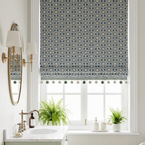 May include: A Roman shade featuring a repeating blue and white floral geometric pattern with a row of small, alternating light gray and sage green pom-pom trim along the bottom edge, installed over a window with white frames and muntins.
