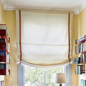 Può includere: Una tenda romana bianca con bordatura arancione è appesa a una finestra. La tenda è tirata su, rivelando una vista di alberi all'esterno. La finestra è incorniciata da una cornice bianca e un muro giallo. Mensole per libri con staffe in metallo rosso sono su entrambi i lati della finestra.