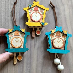May include: Three miniature wooden cuckoo clocks in shades of yellow and teal. Each clock features a round clock face with hands, a pendulum, and decorative bird accents. They hang from brown cords.