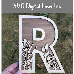 May include: Wooden letter "R" with a floral design and a wooden background. The letter is held by a hand. The text "SVG Digital Laser File" is at the top. The letter is made of light-coloured wood.