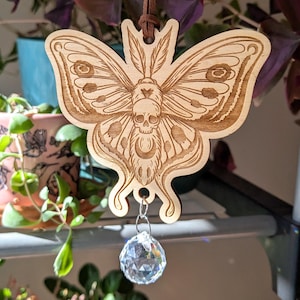 Puede incluir: Un adorno de madera con forma de mariposa con un diseño de calavera, colgado de un cordón marrón. La mariposa tiene patrones de alas detallados y una bola de cristal transparente. El adorno cuelga frente a plantas.