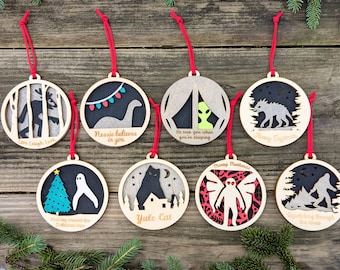 May include: A collection of eight wooden Christmas ornaments with red hanging strings. Each ornament features a unique design, including a Bigfoot, Nessie, an alien, a wolf, a ghost, a cat, a Mothman, and a Bigfoot in the snow.