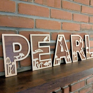 May include: Wooden letters spelling "PEARL" with a light wood border and a dark wood interior. Each letter features laser-cut designs of mushrooms, animals, and other nature-themed elements. The letters are displayed on a dark wooden shelf against a brick wall.