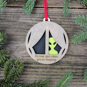 May include: Wooden Christmas ornament with a laser-cut design featuring an alien figure in neon green. The ornament has a gray finish with a red hanging cord and the text "He sees you when you're sleeping".