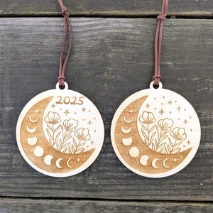 May include: Two round wooden ornaments with a moon phase design, flowers, and the year 2025 etched into the wood. Each ornament has a brown leather cord for hanging. The ornaments are light brown in color.