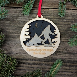May include: Wooden Bigfoot ornament with a red ribbon for hanging. The ornament features a cutout design of Bigfoot walking through snow, with a forest and night sky background. Text reads "Squatching through the snow."