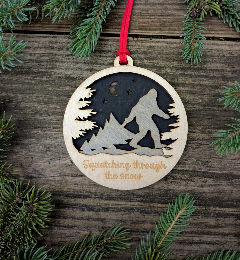 May include: Wooden Bigfoot ornament with a red ribbon for hanging. The ornament features a cutout design of Bigfoot walking through snow, with a forest and night sky background. Text reads "Squatching through the snow."
