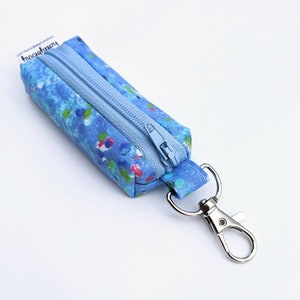 Puede incluir: Pequeño estuche rectangular azul con cierre de cremallera y mosquetón plateado. El estuche está decorado con un patrón de acuarela en azul, verde y rojo. El texto "homeberry" está impreso en una pequeña etiqueta.