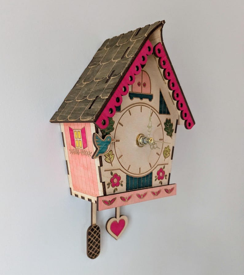 May include: A pink and brown wooden cuckoo clock shaped like a house with a bird and flowers painted on it. The clock has a pendulum with a heart-shaped weight.